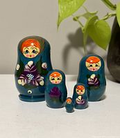Five vintage Russian matryoshki nesting dolls in a row from largest to smallest on a white surface with green plant in background.