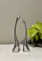 Front and side view of two sleek silver bird-like sculptures on white surface next to green plant