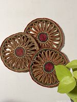 Three round wicker trivets displayed overlapping on a white surface, showing their intricate woven patterns and red and green centers.
