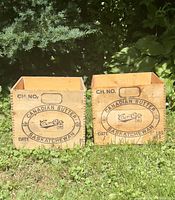 Two wooden crates with printed Canadian Butter Saskatchewen logos and Co-Op branding, positioned on grass with foliage background, showing front and top views.