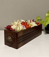 Angle view showing rectangular dark wooden planter with carved detail and metal ring at the end, filled with multicolored artificial flowers including white, red, orange, and purple.