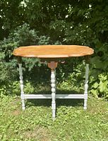 Full view of vintage wooden table with light brown carved wooden oval top and white painted turned legs standing on grass with foliage backdrop.