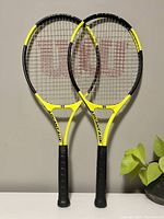 Front view of two Wilson tennis rackets showing black and yellow frames and string pattern with large red Wilson 'W'.