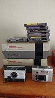 Two Nintendo NES consoles stacked with nine NES game cartridges on top. Kodak Brownie 134 film camera and HP digital camera in front.