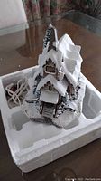 Snow-covered small church sculpture with steeple, showing front entrance and snow on roof and surrounding ground. Includes power cord likely for lighting.