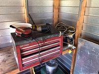 Red Craftsman four drawer tool box with tools on top and coiled cables behind it, located in a metal shed corner.