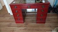Full front view of the wooden desk showing red paint, drawers with brass oval handles, and inlaid top surface under glass.