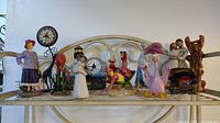 Shelf with diverse figurines featuring vintage woman, flamingos, Willow Tree human figures, and two small round clocks on metal stands.