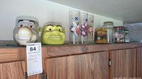 Photo showing Shrek and Donkey ceramic mugs, two Celebrity Cruise glasses, and multiple glass storage jars and bowl on a wood shelf.