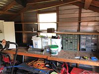 Wide shot of shed shelf with three white buckets, a green and blue plastic small parts organizer filled with hardware, alongside other tools and items on shelf.