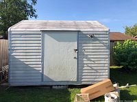 Front view of the Smithbilt metal shed with closed 4 ft door, showing weathered gray siding, metal handle and lock on door, and a security camera or light mounted above the door.