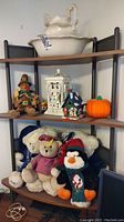 Three-tier shelf with vintage white ceramic wash basin and pitcher on top shelf, decorative lantern, Christmas house, small pumpkin and scarecrow on middle shelf and various stuffed animals on bottom shelf.