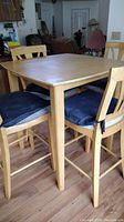 Photo showing square wooden table with four matching wooden chairs with black cushions, all in light wood finish, table surface stained
