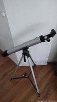 Full view of the silver Vivitar refractor telescope mounted on the silver tripod stand with black fixtures on hardwood floor near wall.
