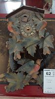 Front view of the cuckoo clock showing carved wooden leaves, three birds, Roman numeral clock face without hands, and parts of the chain and pinecone weights.