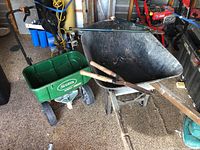 Wide angle showing iron wheelbarrow with deep basin, Scotts green fertilizer spreader, and pruners on top inside garage setting with other tools visible.
