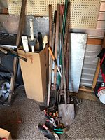 Photo showing long-handled yard tools including shovels, rakes, and others standing next to a cardboard box containing smaller tools.