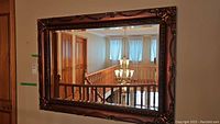 Full frontal view of the large rectangular wall mirror showing the reflection of a stair railing and chandelier. The frame is ornately carved with a raised pattern and finished in bronze/gold tones.