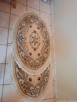 Top view of two oval wool rugs with floral designs and white fringes placed on tiled floor