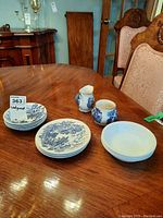Three Wedgwood blue and white side plates stacked with four Wedgwood blue and white bowls adjacent, plus a blue and white English creamer pitcher and sugar bowl placed beside white Corelle bowls on wooden table.