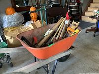 Orange metal wheelbarrow filled with assorted garden and hand tools placed inside a garage or indoor storage area with pumpkins and other items in background.
