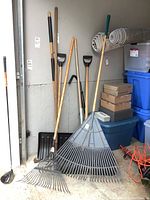 Wide view of six yard tools leaning against a garage wall including two fan rakes, two snow shovels, a weed puller, and a small rake.