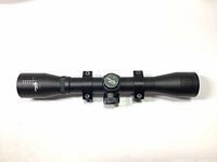 Side view of black Excalibur rifle scope mounted in two black scope rings laid flat on white surface.