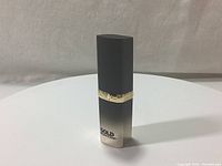 Front side view of lipstick showing gradient black to gold with gold band and text.
