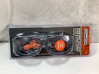 Front view of sealed packaging showing clear lens shooting glasses with black frame and orange foam ear plugs inside transparent plastic blister pack.