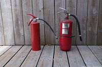 Two red ABC powder fire extinguishers standing upright on wood floor with a wooden fence background.