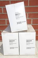 Three white boxes of HP Q6470A (501A) black toner cartridges stacked against a brick wall. Each box is labeled with the part number and NuPrint identification code.
