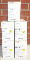 Five boxed yellow toner cartridges, CE412A, stacked in front of a brick wall.