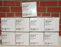 Nine white boxes of NuPrint replacement toner cartridges labeled CF211A (HP 131A) stacked against a brick wall.