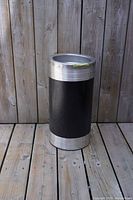 Full view of vintage cylindrical Mid Century Modern floor ashtray with black steel body and silver metal top and bottom edges standing on wooden deck against wood panel background.