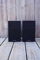 Pair of black Realistic Minimus 26 bookshelf speakers front view on wooden surface with fence background.