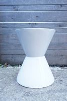 Front view of the white hourglass-shaped pedestal table placed on a rough concrete surface with a wooden fence background.