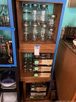 Tall wooden shelf with multiple loose Ball Mason jars of varied sizes and shapes stacked on shelves, with three green Ball brand boxed sets on middle shelf.