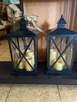 Front view of two black metal lanterns with glass panes and candlesticks inside, one with a gold bow on the handle.