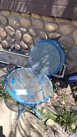 Photo showing two blue round folding metal tables with mesh surfaces placed near a stone wall outdoors on gravel.
