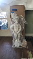 Little boy garden fountain statue wrapped in protective plastic in front of cardboard packaging, side frontal view.