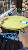Photo of outdoor round painted wood table with lime green top showing some dirt and weathering, surrounded by blue metal framed bench and chair with wooden slats.