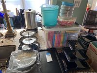 Black plastic three-tiered serving trays alongside turquoise plastic ice bucket and stacks of plastic cups, plates, and napkins on a countertop.