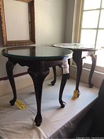 Two Queen Anne style wooden end tables with round glass tops shown side by side on a white draped surface. Both tables have dark wood finish and curved legs.