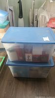 Two stacked blue‐lidded storage bins holding ornament boxes and loose figures
