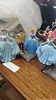 Four princess figurine ornaments on table including three Cinderella in different dresses and one Prince Charming character.