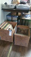 Two cardboard boxes filled with vintage vinyl records placed on a floor beside a table with turntables on top. Visible records include a Luciano Pavarotti album.