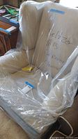 Front view of off white wingback fabric chair covered in clear protective plastic with writing visible through it.