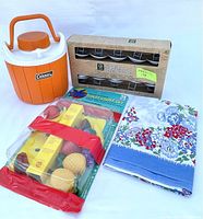 Photo showing all items together: Coleman thermos, box of metal patio string lights, package of fruit-shaped string lights, and folded floral tablecloth.