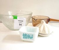 Four kitchen items together: clear floral glass bowl, amber Vision sauce pot, white Pyrex Butterprint refrigerator dish with rooster design, and ceramic garlic roaster.
