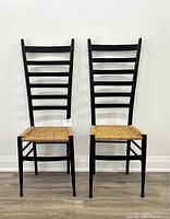 Front view of two black ladder-back chairs with woven rush seats against a white wall